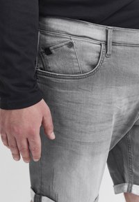 Shorts en denim gris avec une texture usée, des ourlets retroussés et un design de poche subtil. Portés avec une chemise à manches longues noire.