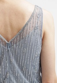 Harmaa helmikirjailtu mekko V-mallisella selällä, jossa on pystysuorat hopeiset yksityiskohdat ja vetoketjusuljenta. Herkkä verkkokangas, jossa on monimutkaisia kuvioita.