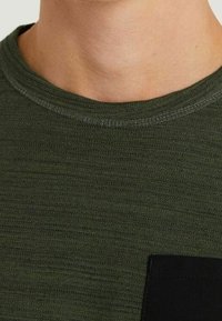 Camiseta de punto de color verde oscuro con textura, cuello redondo y un detalle de bolsillo negro en el lado izquierdo.