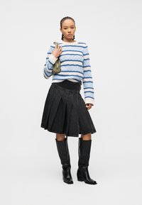 Νεαρό άτομο με κοτσίδες cornrow, φοράει μπλε ριγέ πουλόβερ, μαύρη πλισέ φούστα, μαύρες μπότες μέχρι το γόνατο και κρατά μια μικρή τσάντα σε ελιά πράσινο χρώμα.