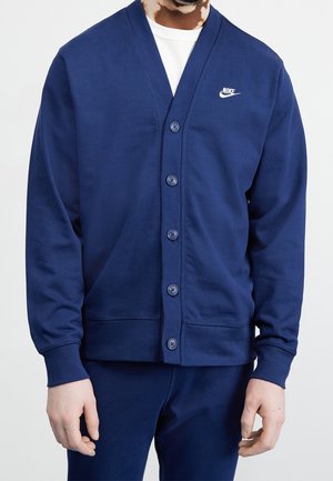 Homme portant un cardigan Nike bleu marine boutonné par-dessus une chemise blanche, assorti à un pantalon bleu marine, debout devant un fond uni.