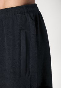 Fekete frottír sort, texturált anyaggal, rugalmas derékrésszel és oldalsó zsebbel. Az anyag lágy és nedvszívó hatásúnak tűnik.