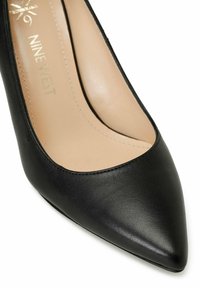 Black leather high heel with a pointed toe, smooth texture, and inner nude lining. Features minimalistic design and no visible hardware.