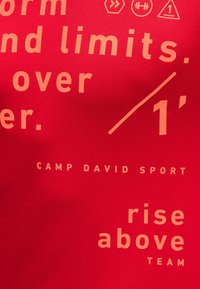 Red fabric T-shirt with large, light beige text featuring phrases "CAMP DAVID SPORT" and "rise above TEAM" in various sizes and styles.