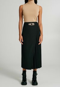 Gonna midi plissè nera dal design elegante, accentuata da una fibbia circolare argentata su una cintura coordinata. Abbinata a un top beige senza maniche e stivali neri.