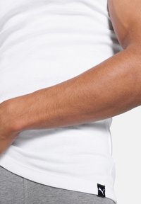 Weißes, tailliertes Tanktop mit glatter Textur, versehen mit einem kleinen schwarzen Puma-Logo-Tag am Saum und grauen Hosen, die unten sichtbar sind.