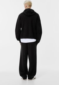 Felpa nera oversize con cappuccio, indossata sopra una camicia bianca, abbinata a pantaloni neri larghi con una tasca posteriore. Texture morbida, design casual.