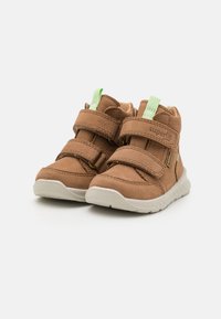 Bottes montantes en cuir marron avec deux bandes Velcro, semelles en caoutchouc blanches et accents verts sur les boucles du talon. Texture lisse et détails cousus.