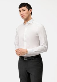 Chemise blanche à manches longues avec patte de boutonnage ; tissu lisse ; col classique ; associée à un pantalon sombre et une ceinture noire.