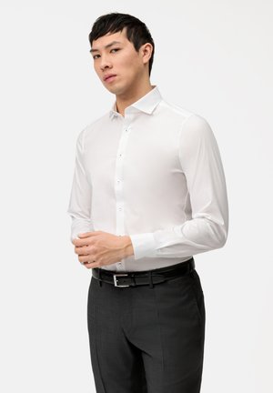 Chemise blanche à manches longues avec patte de boutonnage ; tissu lisse ; col classique ; associée à un pantalon sombre et une ceinture noire.
