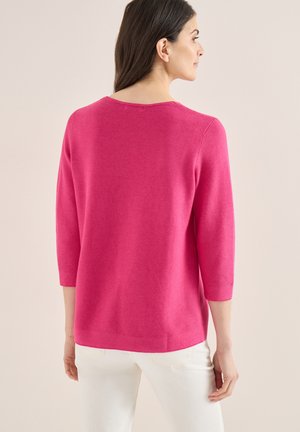 Femme aux cheveux foncés portant un haut à manches longues rose vif et un pantalon blanc, debout de dos sur un fond uni.