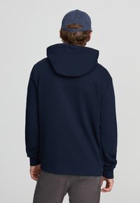 Felpa con cappuccio blu navy in misto cotone, con tasca a marsupio, polsini e orlo a costine. Indossata con pantaloni grigi e un cappellino blu.
