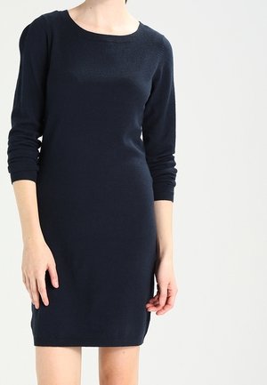 Vrouw draagt een effen, marineblauwe jurk met lange mouwen tot op de knie, staand tegen een lichtgrijze achtergrond.