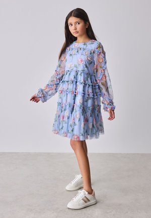 Ragazza con lunghi capelli scuri che indossa un vestito a fiori azzurro chiaro con balze e sneaker bianche, in piedi su un pavimento chiaro davanti a uno sfondo semplice.