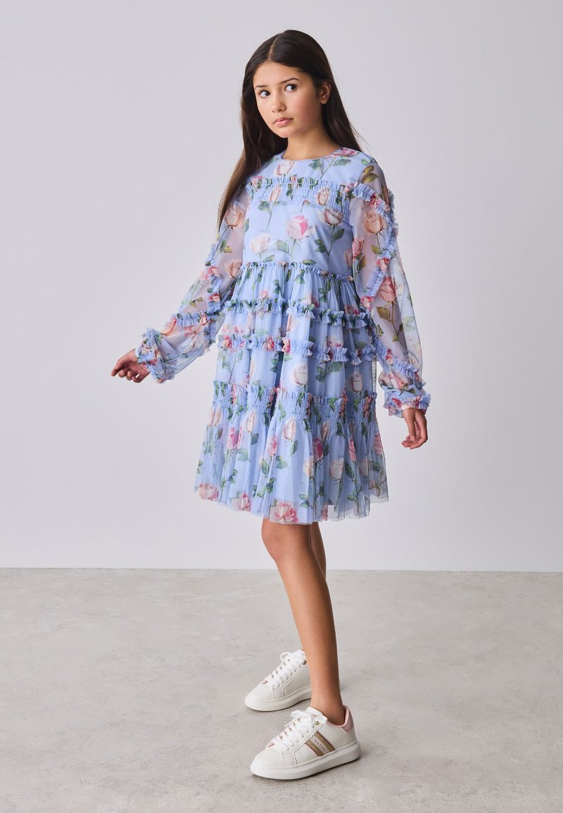 Girl with long dark hair wearing a light blue floral dress with ruffles and white sneakers, standing on a light floor against a plain background.
