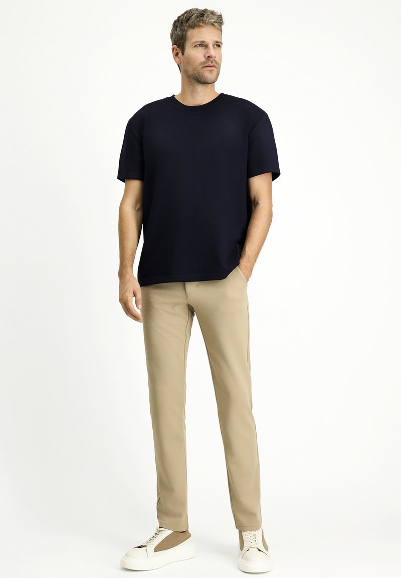 T-shirt blu navy con maniche corte, vestibilità comoda; pantaloni beige in tessuto liscio, abbinati a sneakers bianche con dettagli marroni.