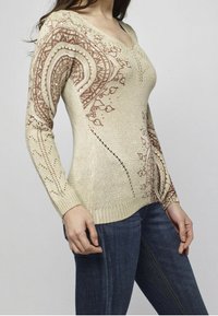 Femme portant un pull beige en tricot avec des motifs bruns complexes sur les manches et le devant, associé à un jean bleu foncé.