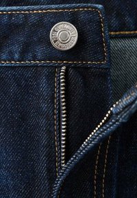 Detailní záběr částečně rozepnutých tmavě modrých džín s viditelným kovovým zipem a stříbrným knoflíkem s logem na pase.