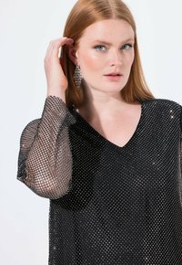 Schwarzes Oberteil mit V-Ausschnitt, verziert mit silbernen Nieten. Durchsichtige Netzärmel mit ausgewähltem Muster. Auffällige Ohrringe ergänzen den Look.