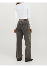 Frau in hochtaillierten, ausgewaschenen schwarzen weit geschnittenen Jeans und einem weißen, langärmeligen, cropped Oberteil, steht vor einem einfarbigen, hellen Hintergrund.