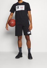 Muž v černém tričku Champion a šortkách drží basketbal, má na sobě černé vzorované ponožky Nike a černé tenisky, stojí na bílém pozadí.