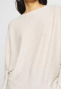 Maglione beige chiaro con una texture morbida, scollatura a barca e vestibilità ampia. Presenta un motivo sottile e cuciture delle spalle abbassate per un maggiore comfort.