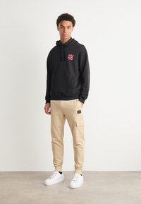 Svart hoodie med röd "HUGO" logotyp, beige cargojoggers med sidofickor, och vita sneakers, som visar upp en avslappnad streetwearstil.