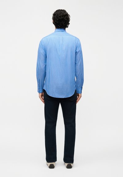 Άνδρας με σγουρά σκουρόχρωμα μαλλιά στέκεται γυρισμένος μπροστά, φορώντας μπλε πουκάμισο με ρίγες και μακριά μανίκια, μαύρο παντελόνι και ανοιχτόχρωμα αθλητικά παπούτσια, πάνω σε λευκό φόντο.