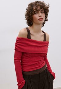 Rotes, geripptes Langarmoberteil mit schulterfreiem Design über einem braunen Tanktop, kombiniert mit dunkler Hose. Mit gerafftem Stoff und enganliegenden Ärmeln.