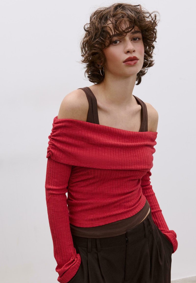 Rotes, geripptes Langarmoberteil mit schulterfreiem Design über einem braunen Tanktop, kombiniert mit dunkler Hose. Mit gerafftem Stoff und enganliegenden Ärmeln.