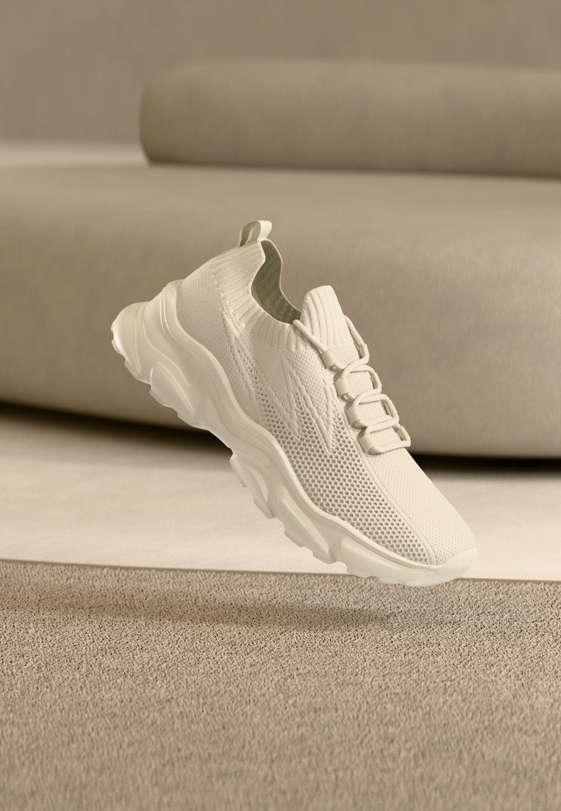 Baskets tricotées beige clair avec semelle épaisse et col montant, montrées en l'air au-dessus d'un tapis beige texturé.