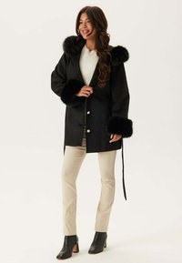Cappotto nero con dettagli in pelliccia sintetica sul cappuccio e sui polsini, caratterizzato da un design su misura e chiusura con bottoni dorati, abbinato a pantaloni beige.