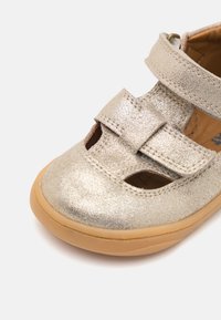 Metallisk sølv sandal med utskjæringsdesign, justerbar stropp og tan gummisåle. Har teksturert overflate og søm detaljer.