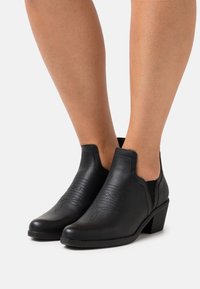 Botas de tobillo de cuero negro con una superficie texturizada, paneles laterales elásticos y un tacón bajo tipo bloque. Diseño de hojas en relieve en la zona de los dedos.