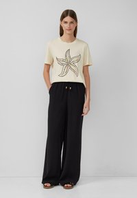 Cremfarbener T-Shirt mit kurzen Ärmeln und einem Seestern-Motiv, kombiniert mit lockeren schwarzen Hosen mit Kordelzug und Holzperlen.