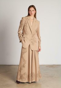 Costum bej cu broderie 3D florală pe jachetă și fustă. Bleaierul croit are rever înclinat și buzunare frontale; fustă lungă, fluidă.