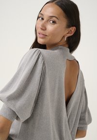 Pull en métal argenté avec des manches bouffantes, un col haut et un grand dos ouvert, reflétant la lumière avec une texture lisse.