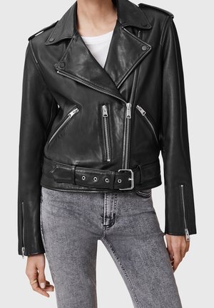 Chaqueta de cuero negra de motorista con cremalleras plateadas y cinturón, llevada sobre una camiseta blanca con vaqueros grises.