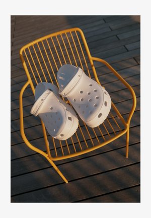 Λευκά Crocs καλοκαιρινά παπούτσια διαθέτουν τρύπες σχεδίασης, επίπεδες σόλες και ρυθμιζόμενους ιμάντες φτέρνας, τοποθετημένα σε μια κίτρινη καρέκλα από σύρμα πάνω σε ξύλινο δάπεδο.