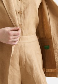 Písečný lněný blejzr s pěti dřevěnými knoflíky, přizpůsobený střih a jemná textura; obsahuje vnitřní štítek v zelené barvě.