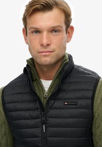 Chaleco acolchado negro sobre un suéter de punto verde, con cremallera frontal y un parche con logo en el lado izquierdo del pecho. Textura suave y diseño ligero.