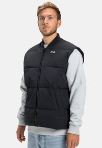 Černá prošívaná vesta s prošitým vzorem, předním zipem a žebrovaným límcem. Nnošená přes světle šedou mikinu s výšivkou loga.