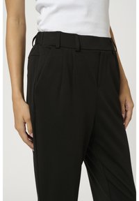 Femme portant un pantalon noir taille haute et un haut blanc sans manches, les mains détendues le long du corps, sur un fond uni.