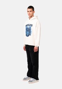 Felpa crema con design grafico blu, jeans neri strappati e sneakers grigie con strisce bianche. Il modello è in profilo su uno sfondo chiaro.