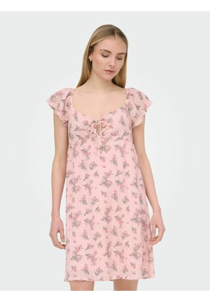 Žena v krátkych ružových kvetovaných šatách s volánikovými rukávmi a detailom viazania na výstrihu, pozerá sa dole s uvoľnenými rukami pri bokoch.