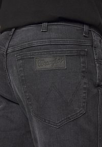 Jeans de mezclilla negra con un bolsillo trasero rectangular que lleva un parche logo "Wrangler" elevado. Las costuras incluyen icónicas costuras en ángulo.