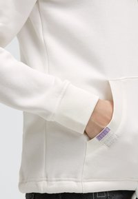 Hand in der Tasche eines weißen Sweatshirts mit geripptem Bündchen und lila Etikett, welches die strukturierte Stoffoberfläche und Nähdetails zeigt.