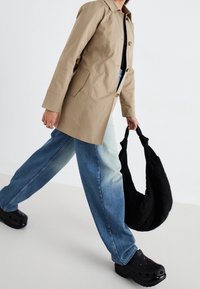 Beżowy płaszcz trench z guzikami, oversized niebieskie dżinsy, czarna pikowana torebka na ramię oraz czarne chodaki z otworami wentylacyjnymi.