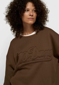 Sweatshirt marron avec texte brodé "10 Days". Le modèle présente une coupe décontractée et un tissu texturé avec un léger éclat.