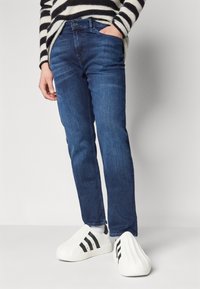 Jeans negre din denim cu o croială dreaptă, având un buzunar frontal și o textură uzată, asortați cu pantofi slip-on albi cu dungi negre.